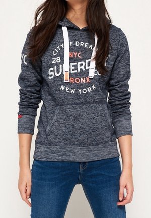 Person wearing a dark gray hooded sweatshirt with white drawstrings and NYC-themed text, paired with blue jeans.