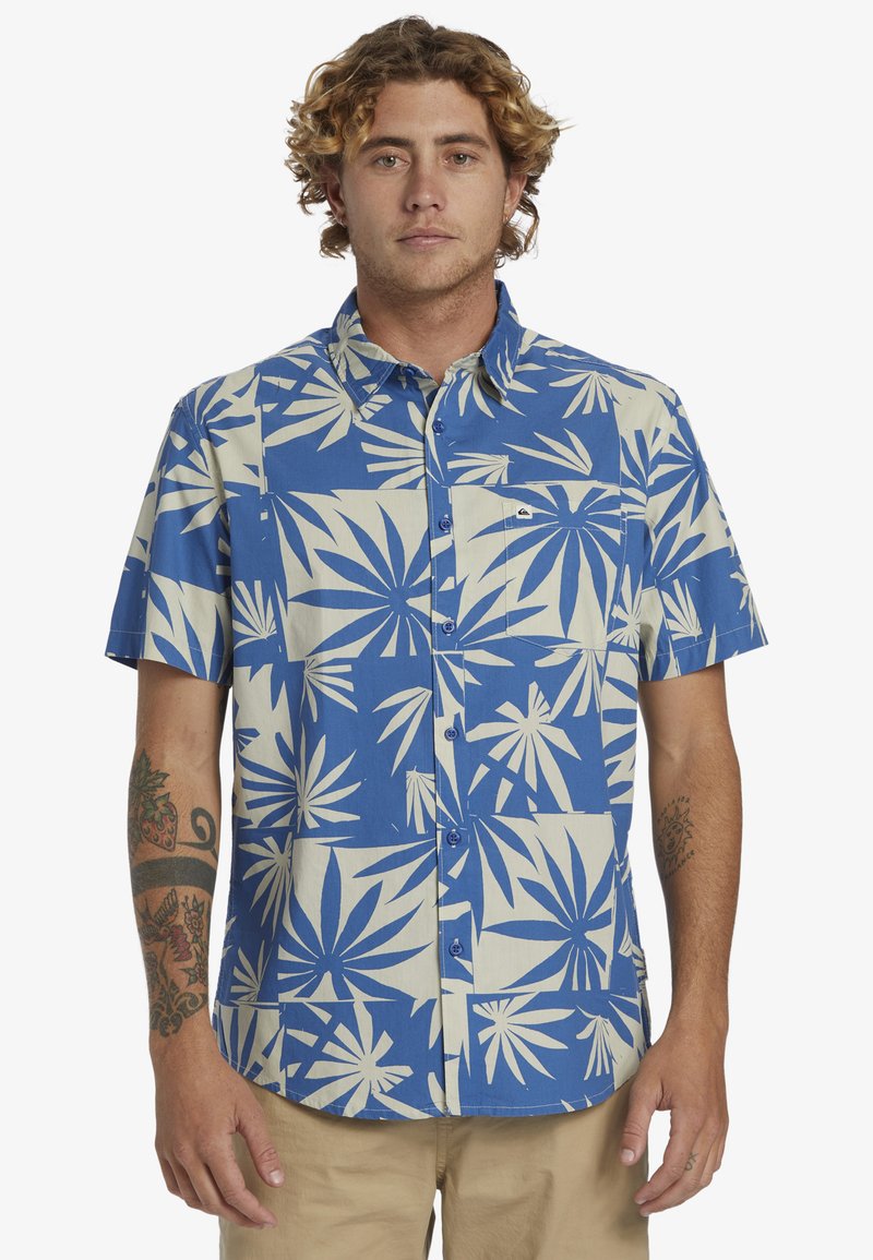 Camisa de botones de manga corta con un patrón de hojas en azul y crema, hecha de algodón, con un bolsillo de parche y botones azules.