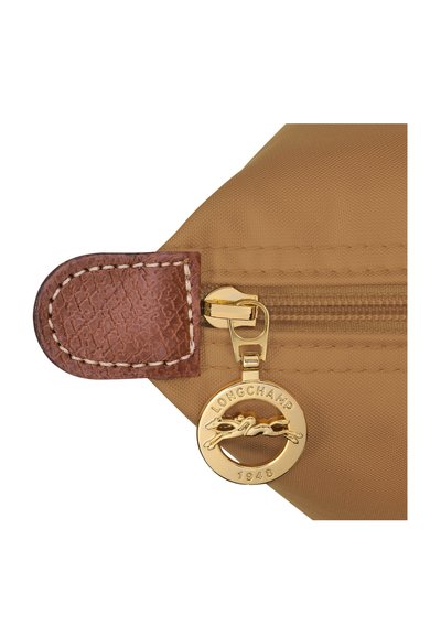 Die tanfarbene Nylon-Tasche verfügt über eine braune, strukturierte Lederlasche und einen goldenen Metallschieber mit einem Logo-Anhänger. Steppnähte sind auf der Lasche zu sehen.