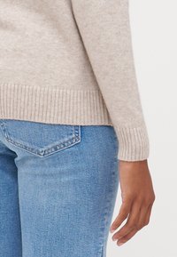 Pullover en tricot beige avec des poignets côtelés, associé à un jean bleu clair. Accent sur la texture douce et la coupe décontractée.