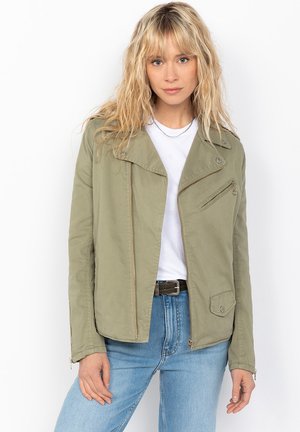Giacca verde oliva con chiusura a zip, tasca inclinata sul petto e spalline. Il tessuto sembra liscio; presenta un design strutturato e corto.