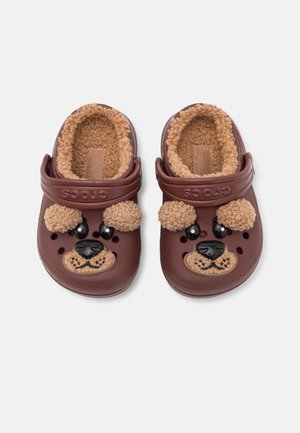 Zuecos marrones sin cordones con forro beige peludo y decoraciones de cara de oso, incluyendo orejas, ojos, nariz y boca en la parte delantera.