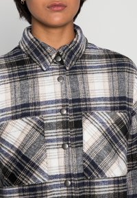 Flanellskjorta i marinblå, svart och vit rutigt mönster; har en kragenek, bröstficka och silverfärgade tryckknappar.