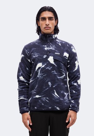 Junger Mann trägt einen marineblauen und weißen gemusterten Fleece-Pullover mit hohem Kragen und geknöpfter Brusttasche und steht vor einem einfarbigen Hintergrund.