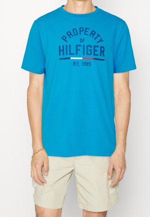 Mann trägt ein hellblaues T-Shirt mit dem Aufdruck „Property of Hilfiger N.Y. 1985“ und beige Cargo-Shorts, steht mit entspannten Armen.