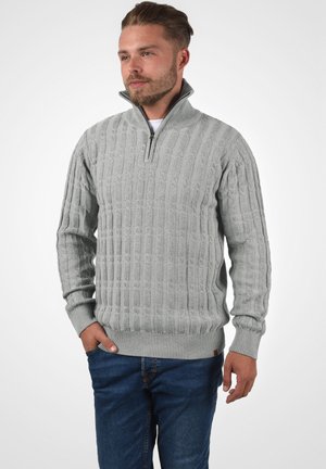 Helehall hall sweater, millel on lukuga kaelus, tekstuuriga nöörkudumine ning soonikud varrukatel ja allservas, koos siniste teksadega.