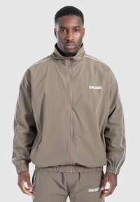Giacca a vento marrone con colletto alto, strisce accentate bianche e chiusura con zip. Presenta un elastico in vita e il logo sul petto.