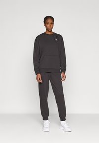 Μαύρο σετ πουλόβερ και παντελόνι, κατασκευασμένο από μαλακό ύφασμα. Διαθέτει τσέπη καγκουρό και μικρό λευκό λογότυπο στο στήθος. Φοριέται με λευκά αθλητικά παπούτσια.