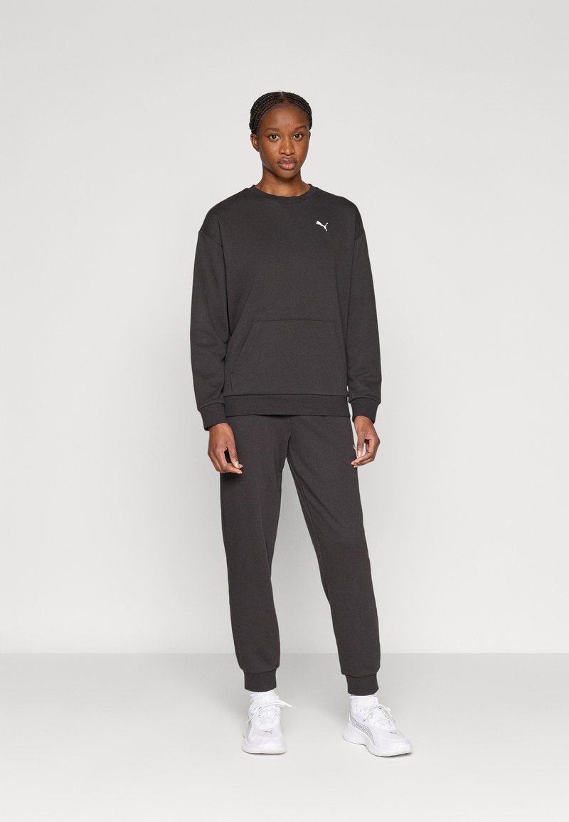 Μαύρο σετ πουλόβερ και παντελόνι, κατασκευασμένο από μαλακό ύφασμα. Διαθέτει τσέπη καγκουρό και μικρό λευκό λογότυπο στο στήθος. Φοριέται με λευκά αθλητικά παπούτσια.