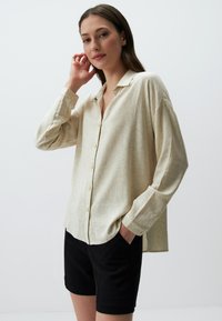 Crème-kleurig, longsleeve overhemd met een subtiel bloemenpatroon en een losse fit. G gedragen met zwarte shorts. Glad textuur.