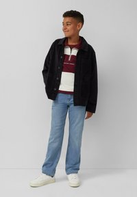 Schwarze Cordjacke mit Knöpfen, kombiniert mit einem bordeauxrot-weiß gestreiften Pullover, hellblauen Jeans und weißen Sneakers.