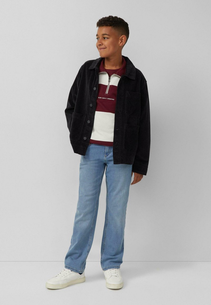 Schwarze Cordjacke mit Knöpfen, kombiniert mit einem bordeauxrot-weiß gestreiften Pullover, hellblauen Jeans und weißen Sneakers.