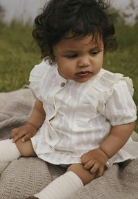 Vestido blanco con volantes y mangas cortas, con sutiles rayas rosas y un botón de madera; niña que lleva calcetines de lana color crema, sentada sobre una manta texturizada.