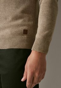 Beige knit sweater with ribbed cuffs and hem, featuring a small dark leather logo patch on the side. Worn with dark green trousers.