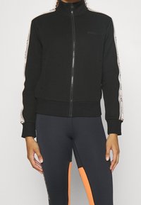 Mujer vestida con una chaqueta negra con cremallera y franjas blancas con patrón en las mangas, y leggings oscuros con paneles verticales naranjas.