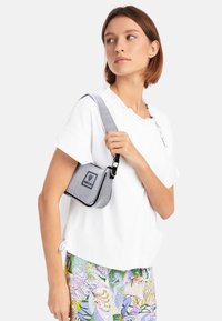 Gestreifte Tasche in Schwarz und Weiß mit abgerundeter Form und verstellbarem Tragegurt, kombiniert mit einem weißen Oberteil mit Rüschenausschnitt und gemusterten Hosen.