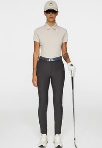 Polo color beige chiaro con collo, pantaloni aderenti grigio scuro, cintura argento con accento blu, scarpe bianche, mazza da golf nella mano destra, guanti sulla sinistra.