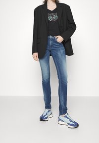 Blazer negro, camiseta negra de cuello en V con un gráfico de tigre, jeans ajustados azules y zapatillas azules con acentos claros y suelas negras.