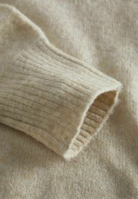 Detalle de un suéter de lana beige que muestra un puño acanalado y una textura suave, con un acabado ligeramente difuso. El tejido de fondo tiene un color claro similar.