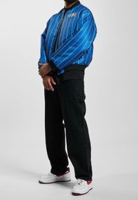 Blauwe satijnen bomberjack met verticale witte strepen en "FUBU"-logo, gecombineerd met zwarte cargobroeken en witte sneakers met rode accenten.