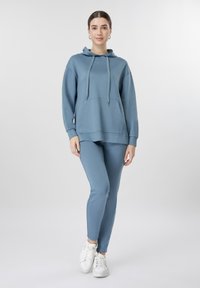 Γυναίκα που στέκεται φορώντας μπλε φούτερ με κουκούλα και ταιριαστά κολάν, με λευκά αθλητικά παπούτσια, μπροστά σε απλό φωτεινό φόντο.