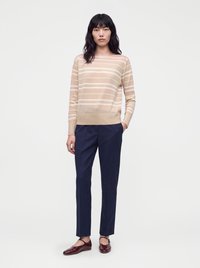 Femme debout portant un pull beige et blanc rayé, un pantalon bleu marine et des chaussures plates rouge foncé avec des brides à la cheville.