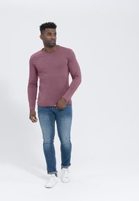 Langärmliges bordeauxrotes Hemd mit rundem Ausschnitt, kombiniert mit eng anliegenden blauen Jeans und weißen Sneakers, vor einem neutralen Hintergrund.