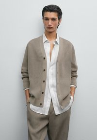 Beige gestrickte Strickjacke mit V-Ausschnitt, Knopfverschluss und gerippten Bündchen, kombiniert mit einem weißen gestreiften Hemd und hellgrauen Nadelstreifenhosen.