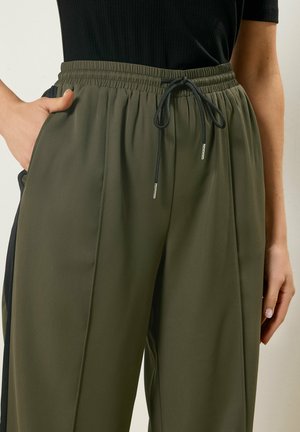 Person iført olivengrønne og sorte elastiske bukser med snøre i taljen, den ene hånd i lommen, kombineret med en sort ribbet top.