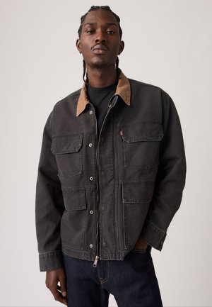 Jeune homme aux cheveux tressés portant une veste noire avec un col beige et plusieurs poches avant, debout devant un fond uni.