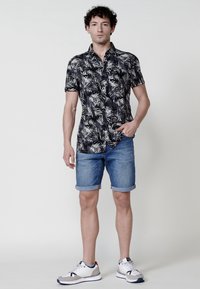 Camisa negra de manga corta con un patrón de hojas blancas, combinada con pantalones cortos de mezclilla azul claro y zapatillas blancas con acentos grises.