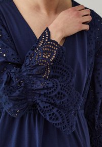 Tummansininen mekko, jossa on brodeeratut pitsihihat. Sisältää monimutkaisia kaiverruksia ja smokkauselementtejä ranteissa. Pehmeä rakenne.