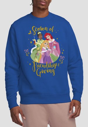 Man wearing blue sweatshirt with Disney princesses Tiana, Ariel, and Rapunzel holding holiday items and text "Season of Friendship & Giving."