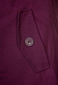 Tessuto viola scuro con un dettaglio della tasca che presenta un bottone argentato. La texture appare liscia, con cuciture visibili lungo i bordi.