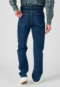 Jeans in denim blu scuro con taglio dritto, dotati di due tasche posteriori e dettagli di cuciture visibili. Abbinati a una camicia a scacchi.