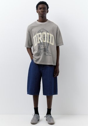 Jeune homme portant un t-shirt beige oversized avec le texte "DROID" et un graphisme de droïde, un short en jean large bleu, des chaussettes noires et des baskets grises.