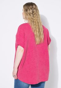 Maglietta over-size a maniche corte di un vivace colore rosa, realizzata in un tessuto texturizzato e intrecciato. Orlo posteriore più lungo e taglio ampio. Jeans blu visibili in basso.