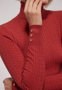 Czerwony sweter z splotem warkoczowym i wysokim kołnierzem, o teksturowanej fakturze. Posiada trzy złote guziki akcentujące na mankiecie dla dodatkowego detalu.