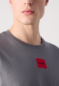 Grå t-shirt med en rektangulær rød lap med ordet "HUGO" i sort. Klassisk crew neck-design med en glat, blød tekstur.