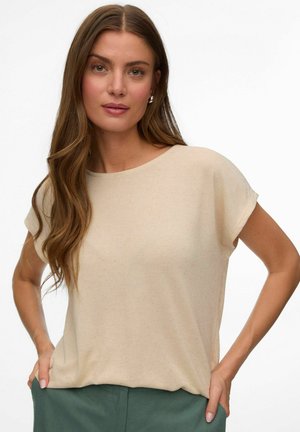 Kvinde med langt brunt hår iført en beige kortærmet top og grønne bukser, stående med hænderne i lommerne mod en ensfarvet baggrund.