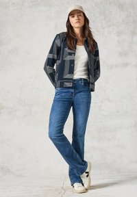 Námořnická vzorovaná bunda s geometrickými liniemi, bílý tílko, modré džíny a bílé tenisky s černými akcenty. Modelka stojí před neutrálním pozadím.