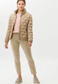 Beige quilted puffer jacket with high collar, featuring a zip front. Worn over a striped shirt and paired with beige pants and light shoes.