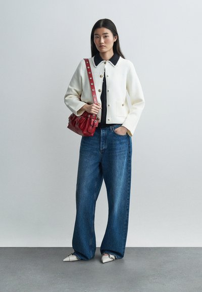 Γυναίκα που φοράει λευκό σακάκι με κουμπιά, σκούρο τοπ με γιακά, φαρδιά μπλε τζιν παντελόνια, λευκά μυτερά παπούτσια, κρατώντας κόκκινη τσάντα ώμου μπροστά σε απλό φόντο.