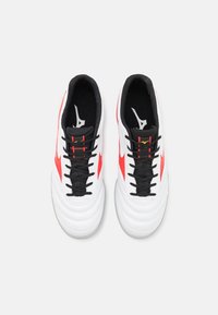 Sneakers bianchi da atletica con lacci e dettagli neri, caratterizzati da dettagli laterali rossi e una parte superiore in pelle testurizzata. Design a profilo basso.
