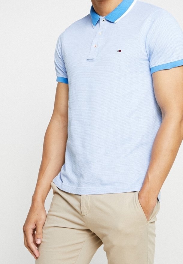 Homme portant un polo bleu clair avec un col et des bords de manches bleus, et un pantalon beige, se tenant avec une main dans la poche contre un fond blanc.