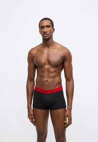 Fekete boxeralsó piros elasztikus derékpánttal, amelyen a márkanév látható. Az anyag simának tűnik, szűk szabású.