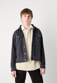 Crna denim jakna s dva prednja džepa, srebrnim gumbima i dizajnom s ovratnikom. Nosiva preko svijetlo bež džempera. Dugi rukavi.
