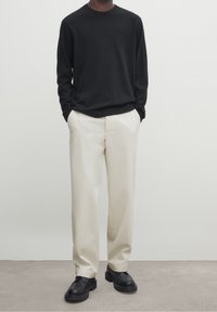 Homme portant une chemise noire à manches longues, un pantalon blanc cassé et des chaussures noires à semelle épaisse, se tenant avec les mains dans les poches devant un fond uni.