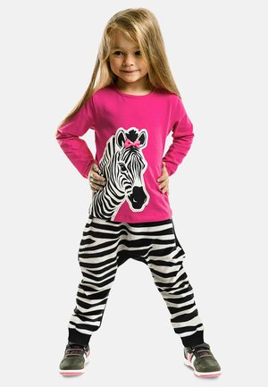 Rosa Langarmshirt mit einem Zebra-Design und einer Schleife, kombiniert mit schwarz-weißen gestreiften Haremhosen. Das Outfit wird mit Sneakers abgerundet.