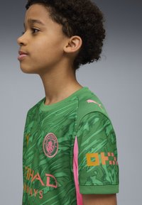 Maglia da calcio verde del Manchester City con un motivo ondulato, dettagli rosa sulle maniche, logo e scritte in giallo e bianco.